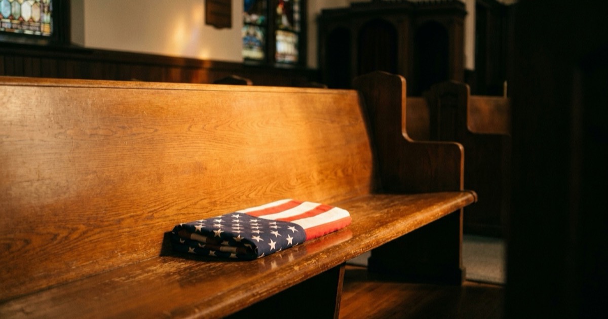 A folded American flag resting alone on an empty wooden church pew in warm afternoon light, the sanctuary dim behind it